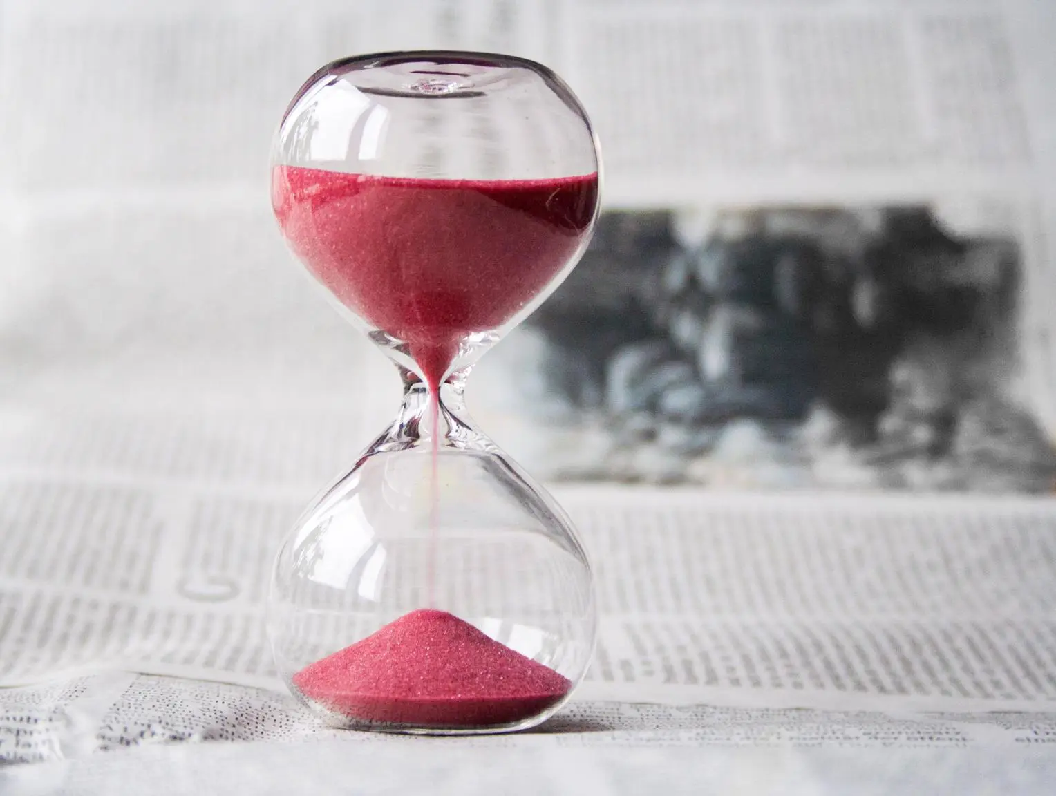 Sablier en verre contenant du sable rouge, représentant le passage du temps, posé sur des pages de journal floues en arrière-plan.